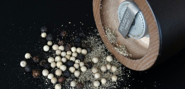 Close-up of a pepper grinder and assorted peppercorns on black surface.