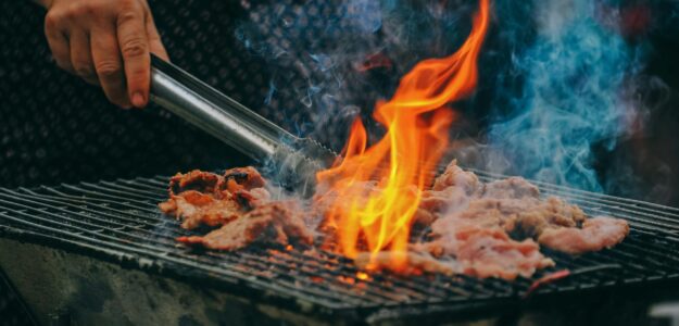 Close-up of grilling meat with vibrant flames and smoky aura, showcasing outdoor cooking.